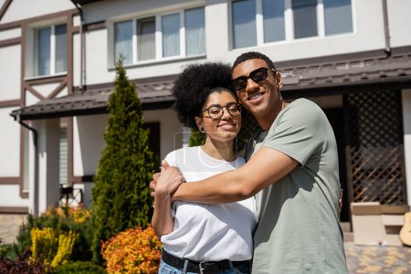 souriant afro-américain en lunettes de soleil étreignant petite amie tout en se tenant près de la nouvelle maison