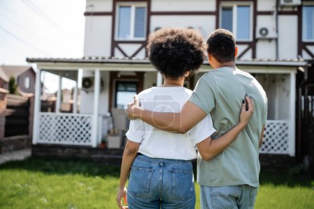 Rückansicht eines afrikanisch-amerikanischen Paares, das sich umarmt, während es in der Nähe eines neuen Hauses im Hintergrund steht