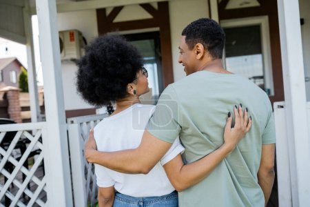sonriente afroamericano pareja abrazándose y mirando el uno al otro cerca de nueva casa