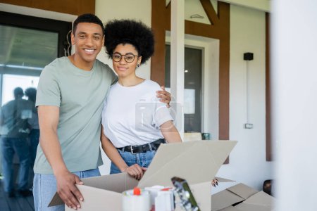 sonriente afroamericano hombre abrazando novia cerca de cajas de cartón y nueva casa durante la reubicación