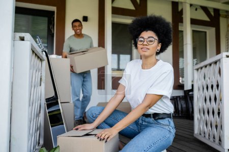 african american woman looking at camera near carton boxes and boyfriend during moving in new house