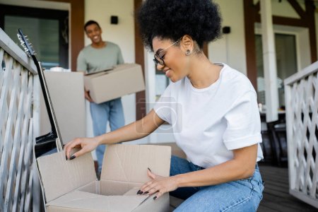 cheerful african american woman unpacking carton box near blurred boyfriend and new house