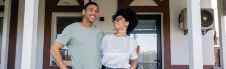 positive african american couple hugging while standing on porch near new house, real estate, banner