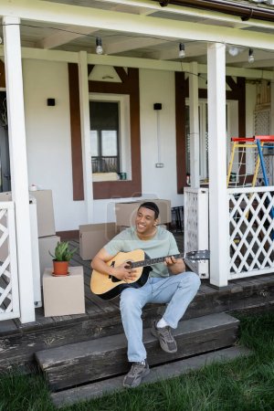 Foto de Alegre afroamericano hombre tocando la guitarra acústica en porche cerca de cajas y nueva casa - Imagen libre de derechos