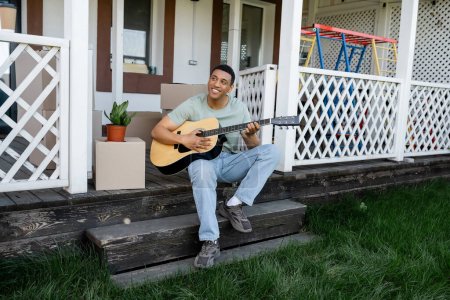 Foto de Alegre afroamericano hombre tocando la guitarra acústica en el porche cerca de cajas de cartón y nueva casa - Imagen libre de derechos