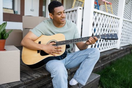 Foto de Alegre afroamericano hombre tocando la guitarra acústica cerca de cajas de cartón y nueva casa - Imagen libre de derechos
