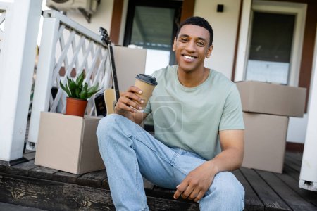 fröhlicher afrikanisch-amerikanischer Mann hält Kaffee in der Nähe von Kartons und neuem Haus im Freien