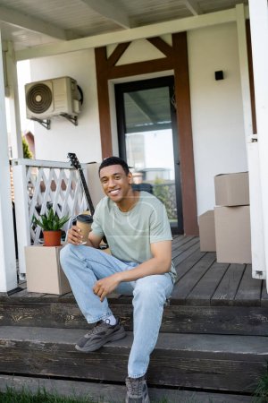 Téléchargez les photos : Souriant homme afro-américain tenant du café pour aller près des paquets sur le porche de la nouvelle maison - en image libre de droit