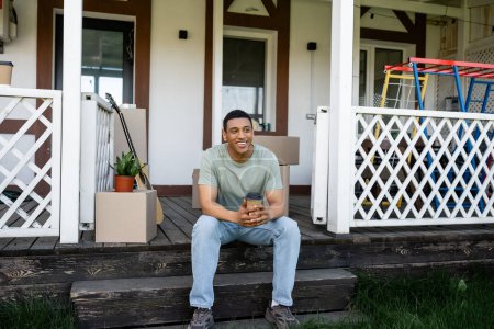 Foto de Sonriente afroamericano hombre sosteniendo el café para llevar mientras está sentado en el porche de la nueva casa - Imagen libre de derechos