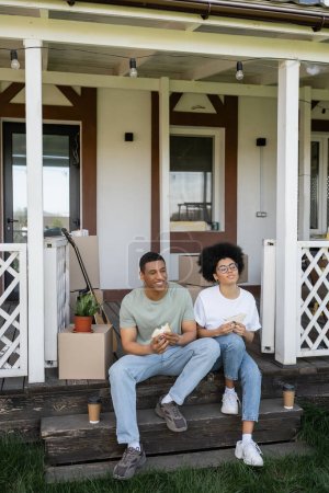 joyful african american couple holding sandwiches near coffee and packages on porch of new house