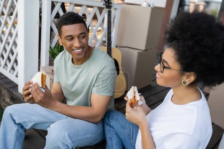 smiling african american man holding sandwich near girlfriend and boxes on porch of new house