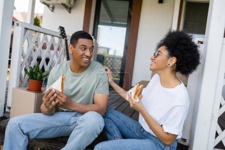 fröhliche afrikanisch-amerikanische Paar reden und halten Sandwiches auf der Veranda des neuen Hauses