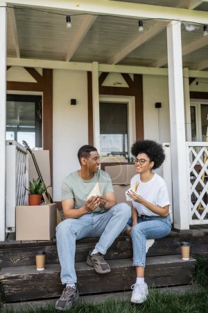 happy african american woman holding sandwich near boyfriend and coffee to go on porch of new house