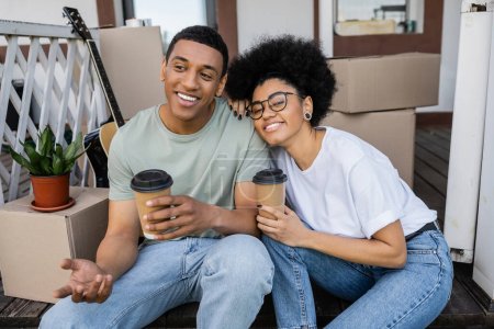 lächelnd afrikanisch-amerikanisches Paar mit Kaffee zum Reden in der Nähe von Boxen auf der Veranda des neuen Hauses
