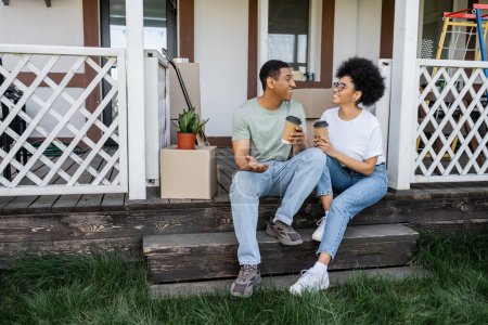 glücklich afrikanisch-amerikanischer Mann mit Kaffee zu gehen im Gespräch mit Freundin in der Nähe von Boxen auf der Veranda des neuen Hauses