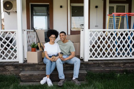 Téléchargez les photos : Sourire couple afro-américain tenant du café pour aller près des paquets sur le porche de la nouvelle maison - en image libre de droit