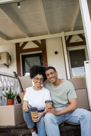 lächelnd afrikanisch-amerikanisches Paar mit Kaffee zu gehen Blick auf die Kamera auf der Veranda des neuen Hauses