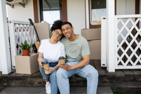 sonriente pareja afroamericana con café para ir abrazando mientras está sentado en el sofá de la nueva casa