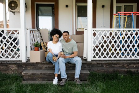 Foto de Alegre africano americano pareja abrazando y celebración café a ir en porche de nueva casa - Imagen libre de derechos