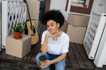 sonriente afroamericana mujer sosteniendo café para ir y sentado cerca de cajas de cartón y nueva casa