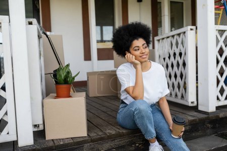 mujer afroamericana complacida sosteniendo café para ir sentado cerca de cajas de cartón y casa nueva