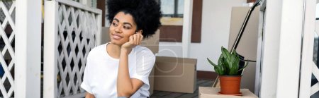 mujer afroamericana soñadora mirando lejos cerca de cajas de cartón en el porche de nueva casa, bienes raíces, pancarta