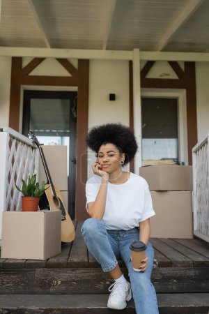 Afrikanische Amerikanerin schaut in die Kamera, während sie Kaffee hält, um auf die Veranda des neuen Hauses zu gehen