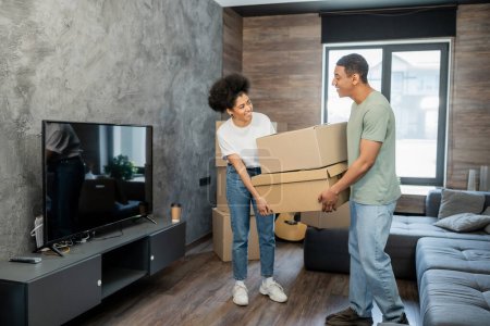 fröhliches afrikanisch-amerikanisches Paar hält Pappkartons in der Nähe von Couch und Fernseher im neuen Wohnzimmer
