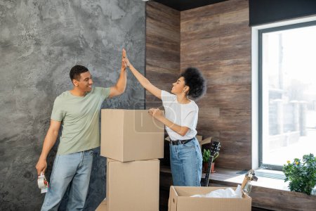heureux couple afro-américain donnant haute cinq près de boîtes en carton dans une nouvelle maison