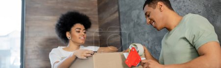 smiling african american man holding adhesive tape near box and girlfriend during moving, banner