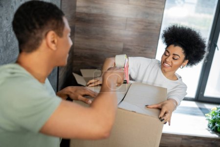 positive african american couple packaging cardboard box with adhesive tape during relocation