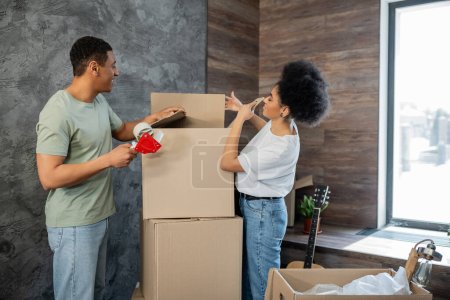 Photo for Side view of happy african american couple with adhesive tape packaging boxes during moving - Royalty Free Image