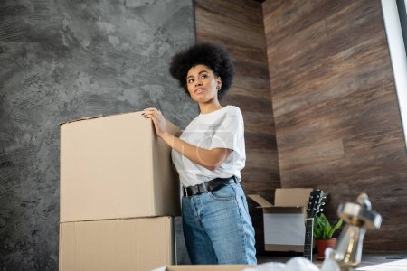 mujer afroamericana de pie cerca de cajas de cartón en la sala de estar en casa nueva