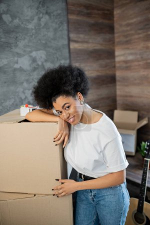 Téléchargez les photos : Femme afro-américaine gaie debout près des boîtes en carton et du ruban adhésif dans le salon - en image libre de droit