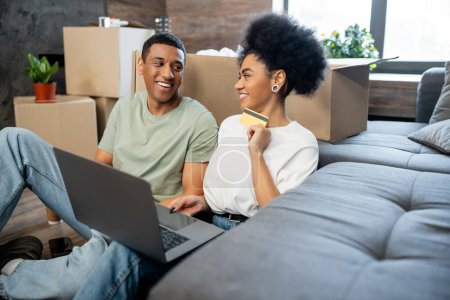 lächelnde Afroamerikanerin mit Laptop und Kreditkarte in der Nähe von Freund und Kisten im neuen Haus