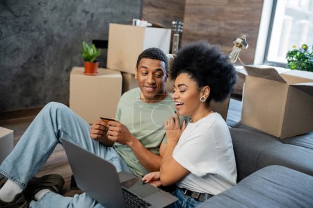 Photo for Carefree african american woman hugging boyfriend with laptop and credit card near boxes at home - Royalty Free Image