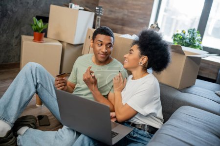 Photo for Joyful african american woman talking to boyfriend during online shopping near boxes in new house - Royalty Free Image