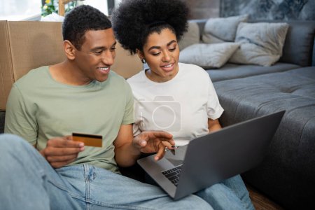 Foto de Pareja afroamericana sonriendo durante las compras en línea cerca de caja de cartón en sala de estar en casa nueva - Imagen libre de derechos