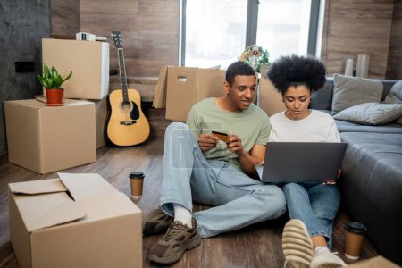 Afrikanisch-amerikanisches Paar beim Online-Shopping in der Nähe von Coffee to go und Kartons im neuen Haus
