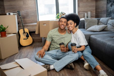 heureux couple afro-américain avec café regardant la caméra près des paquets dans le nouveau salon