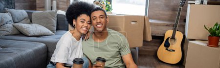 pleased african american couple with looking at camera near cardboard boxes in new home, banner