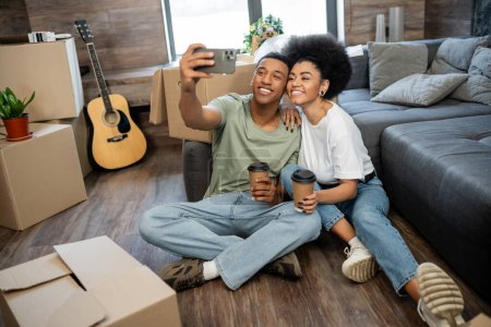 Foto de Alegre pareja afroamericana tomando selfie y sosteniendo café cerca de cajas de cartón en nueva casa - Imagen libre de derechos