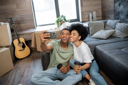heureux couple afro-américain prenant selfie et tenant café près des paquets dans le nouveau salon