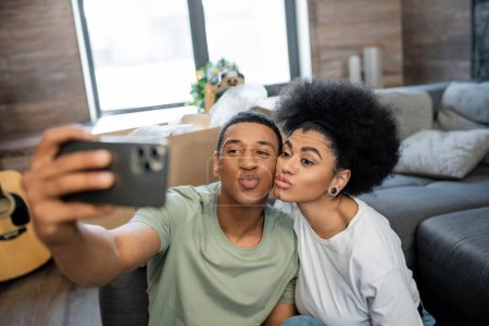 Foto de Pareja afroamericana haciendo pucheros labios mientras toma selfie en el teléfono inteligente en la nueva sala de estar - Imagen libre de derechos