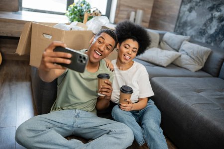 excited african american couple taking selfie and holding coffee in living room in new house