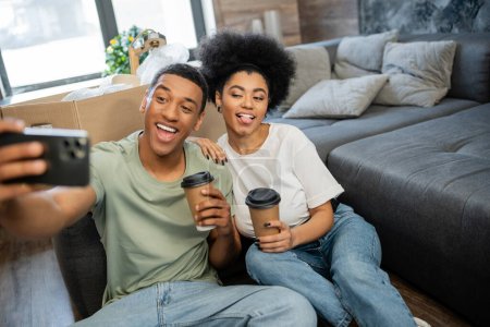 Afrikanisches amerikanisches Paar grimmig, während es ein Selfie mit dem Smartphone macht und Kaffee im neuen Haus hält
