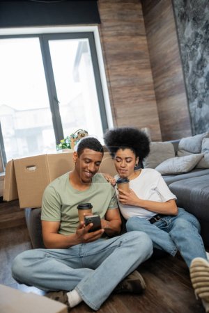 positif couple afro-américain avec café en utilisant un smartphone près des paquets dans la maison neuve