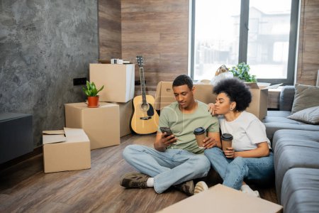 Afrikanisch-amerikanisches Paar benutzt Handy und hält Kaffee in der Nähe von Paketen im neuen Wohnzimmer