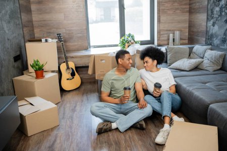 Lächelndes afrikanisch-amerikanisches Paar spricht und hält Kaffee in der Nähe von Schachteln im neuen Wohnzimmer