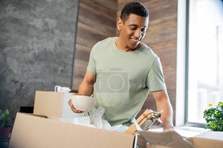 homme afro-américain insouciant tenant des biens près de boîtes en carton dans une maison neuve